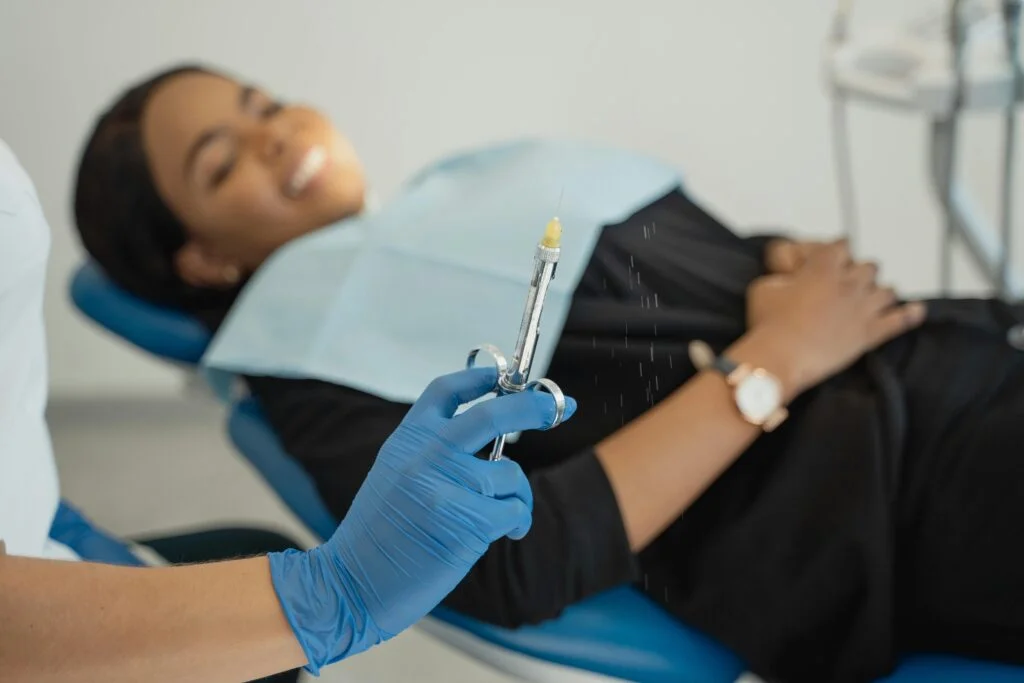 A dentist wearing gloves prepares an injection as a patient smiles in a dental clinic.